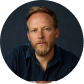 Man with beard and short hair wearing a dark blue shirt against a dark background.